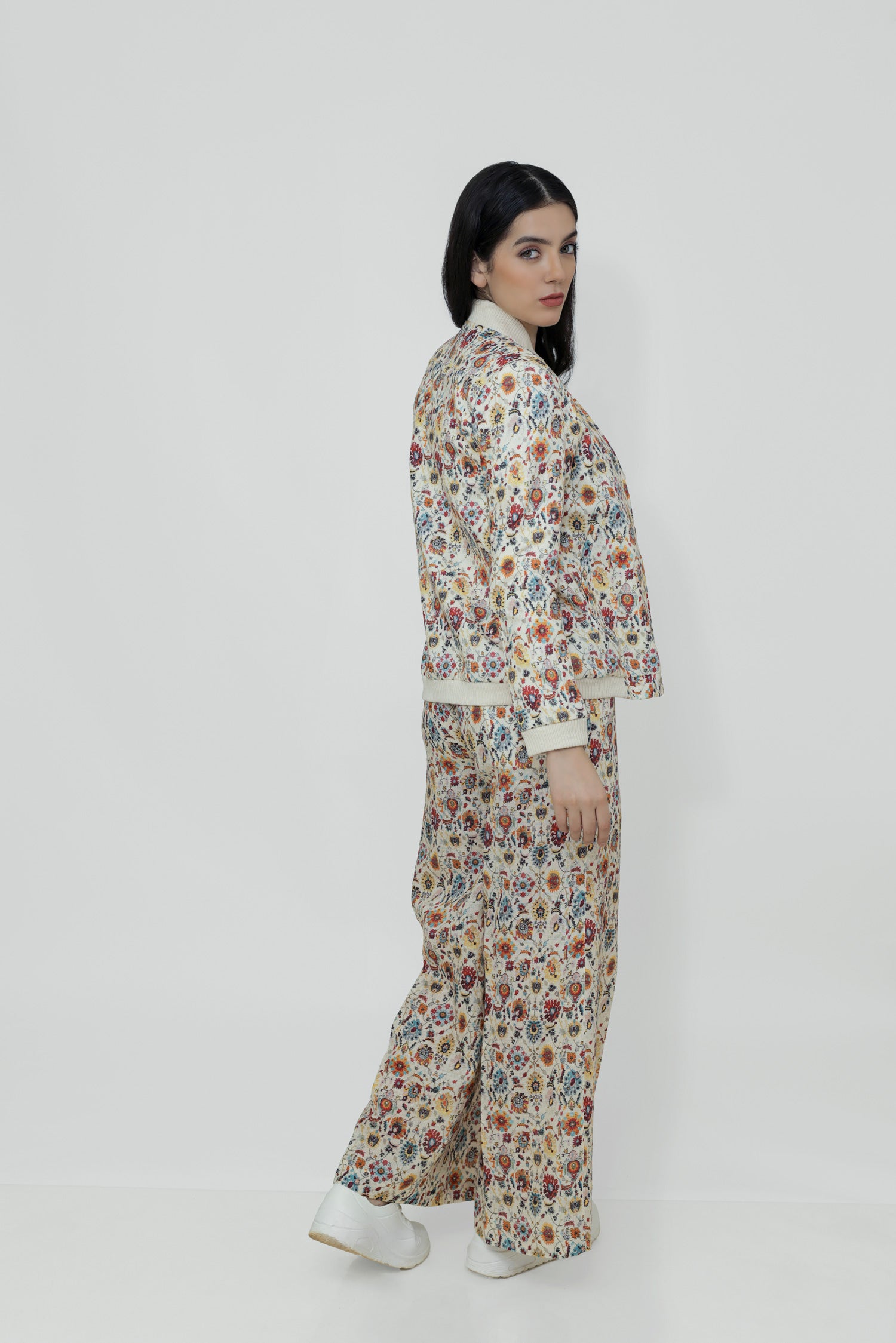 A woman models a floral linen bomber jacket with a collar, featuring a front zipper and ribbed cuffs. She is wearing a white top and white shoes, standing against a plain background.