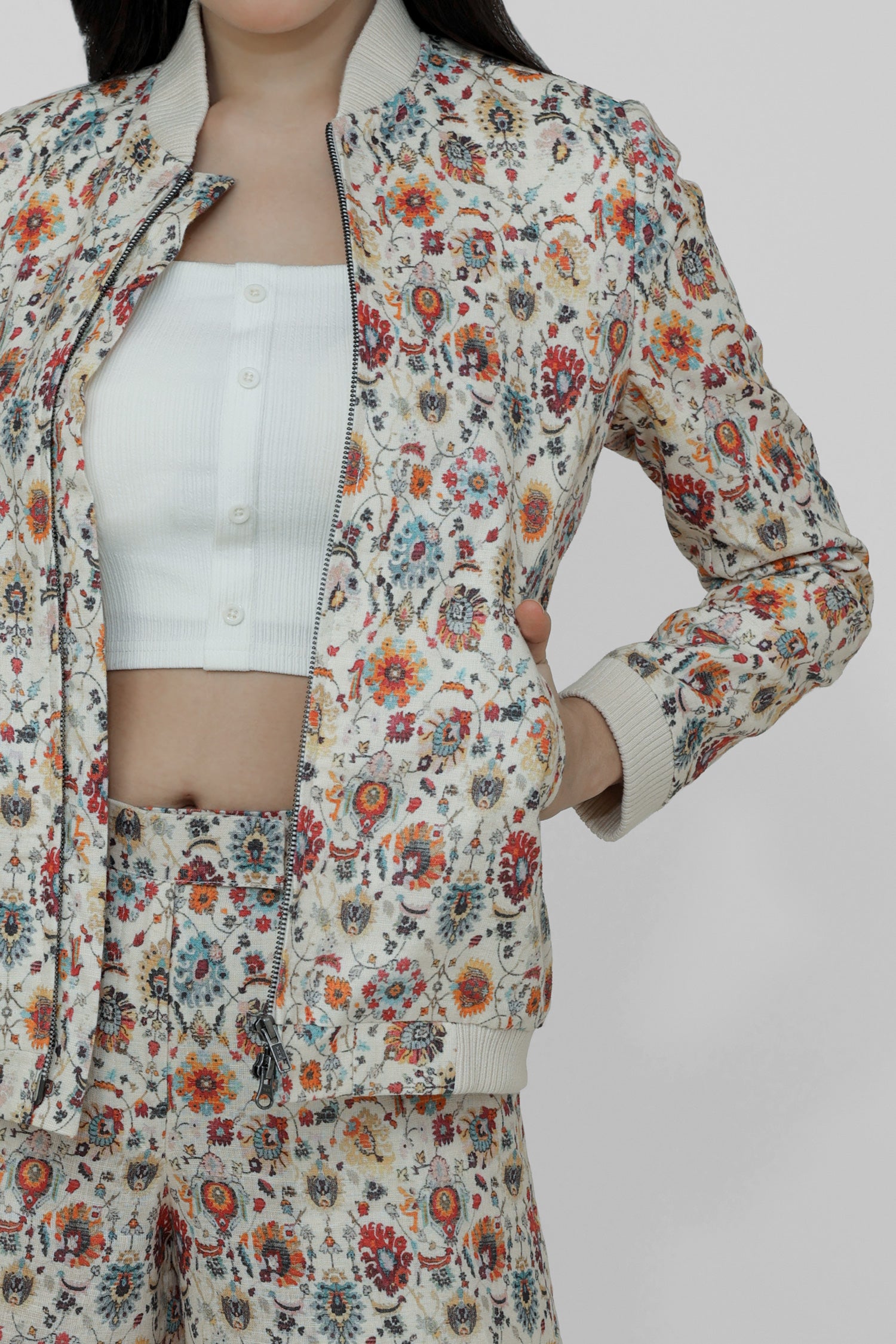 A woman models a floral linen bomber jacket with a collar, featuring a front zipper and ribbed cuffs. She is wearing a white top and white shoes, standing against a plain background.