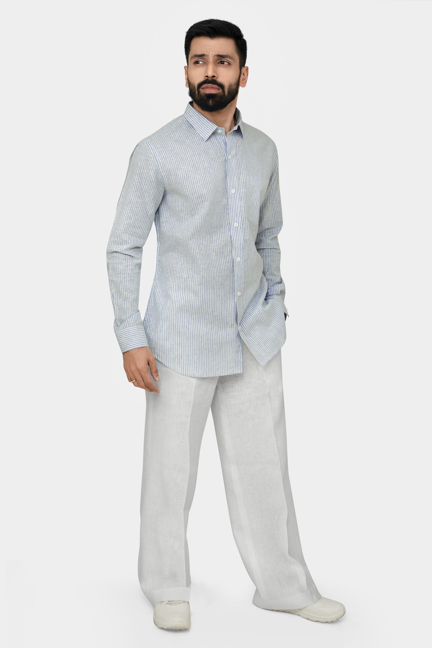 A man standing, wearing a linen shirt with a cut-away collar, long sleeves, and a single-button cuff, paired with light-colored trousers.