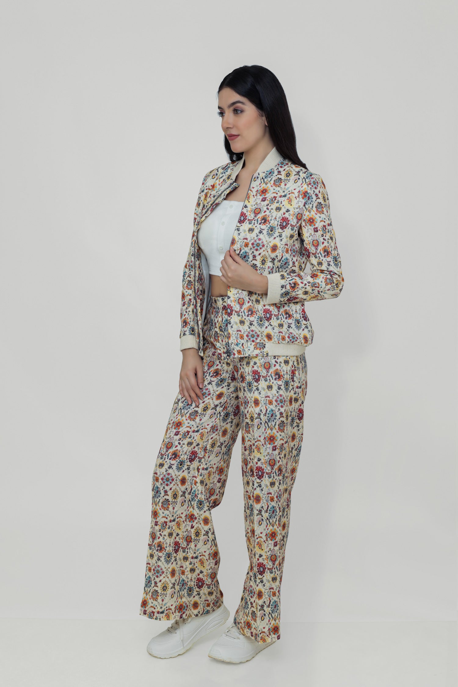 A woman models a floral linen bomber jacket with a collar, featuring a front zipper and ribbed cuffs. She is wearing a white top and white shoes, standing against a plain background.