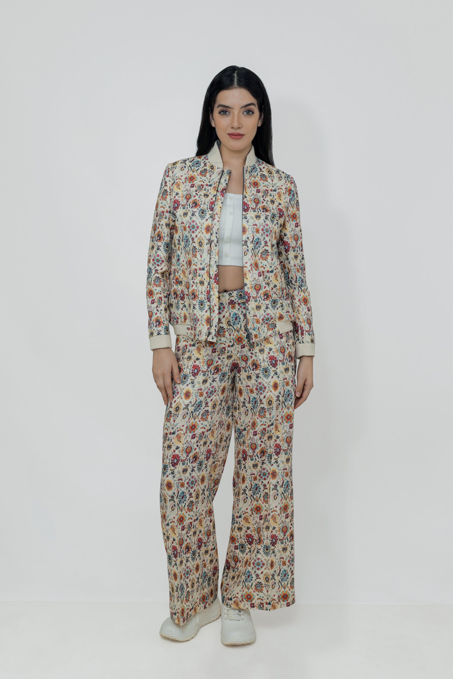 A woman models a floral linen bomber jacket with a collar, featuring a front zipper and ribbed cuffs. She is wearing a white top and white shoes, standing against a plain background.