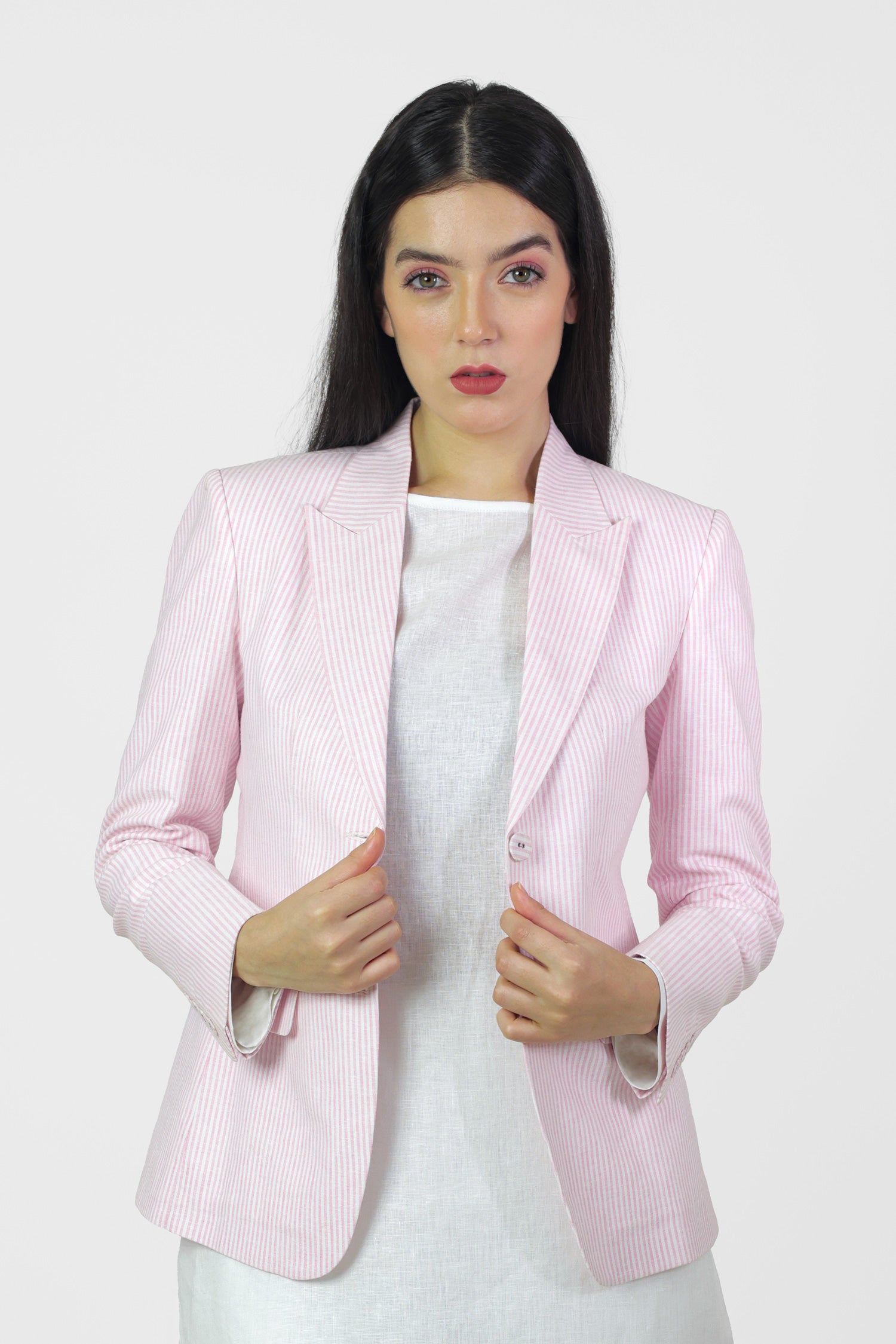 A woman standing outdoors wearing a pink linen blazer with a single-breasted design, peak lapel, and flap pockets. She also has on white trousers and is posing with her hands by her sides.