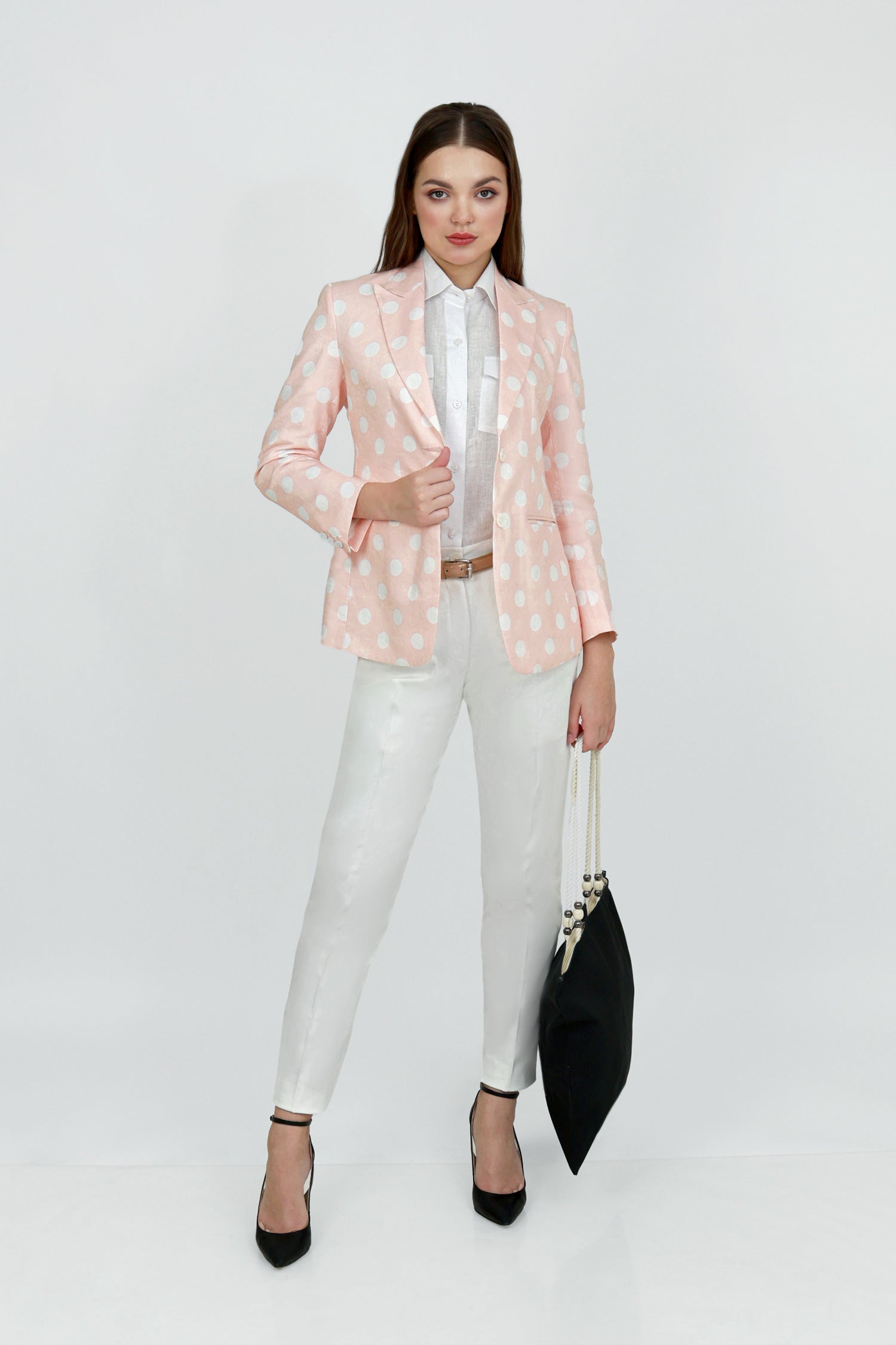 A woman model wearing a peach hemp blazer with a peak lapel, two buttons, and flap pockets, paired with white trousers.