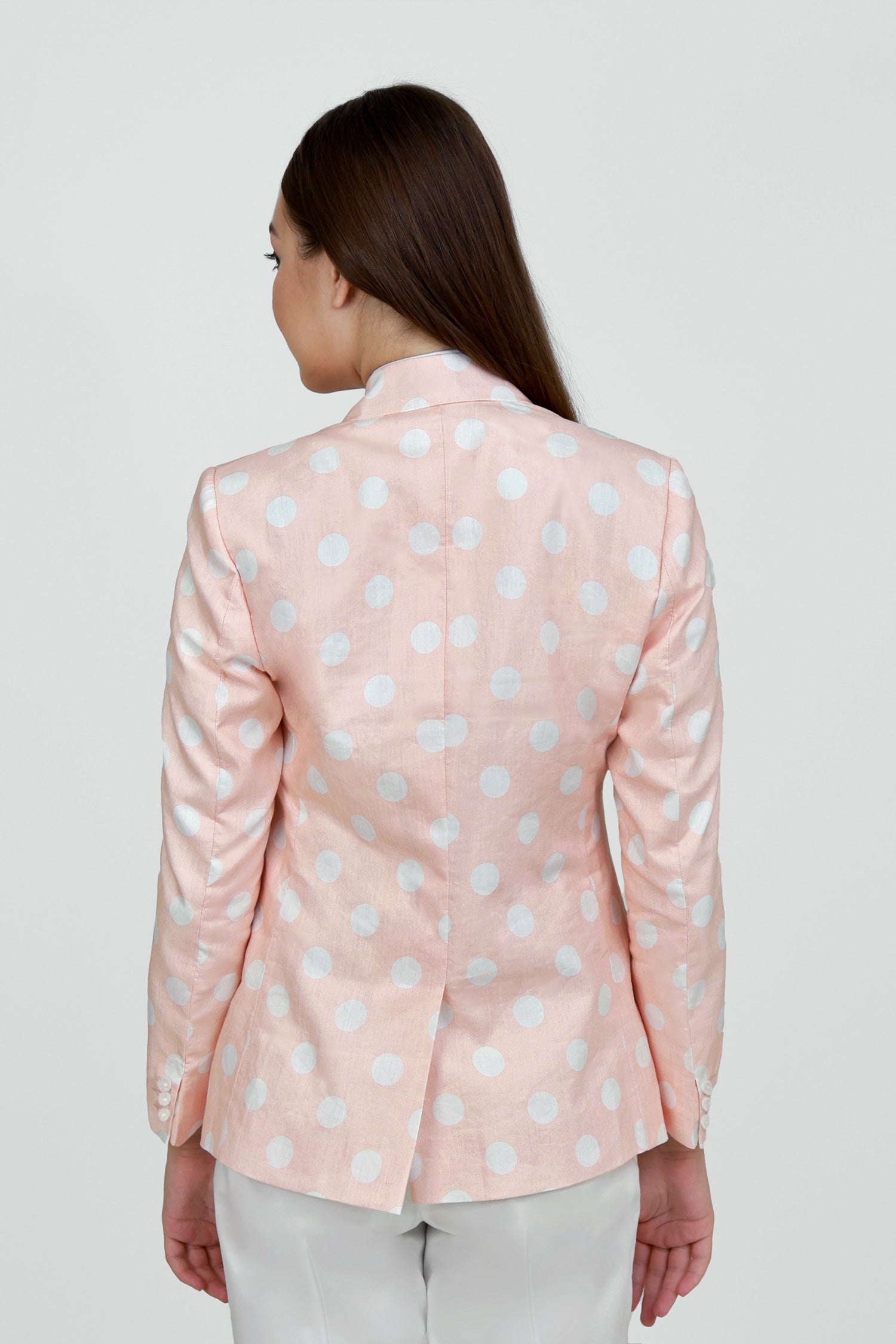 A woman model wearing a peach hemp blazer with a peak lapel, two buttons, and flap pockets, paired with white trousers.