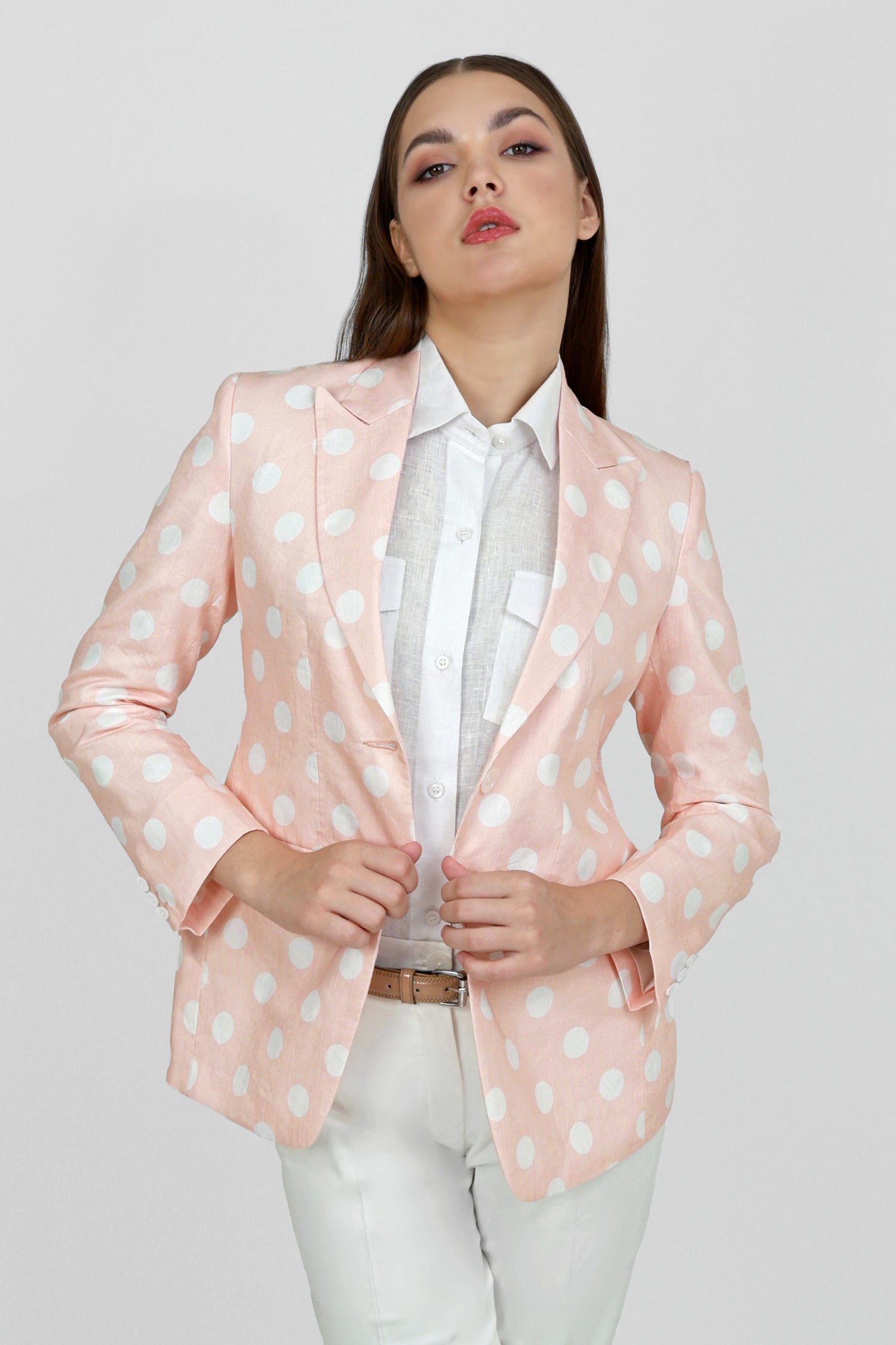 A woman model wearing a peach hemp blazer with a peak lapel, two buttons, and flap pockets, paired with white trousers.