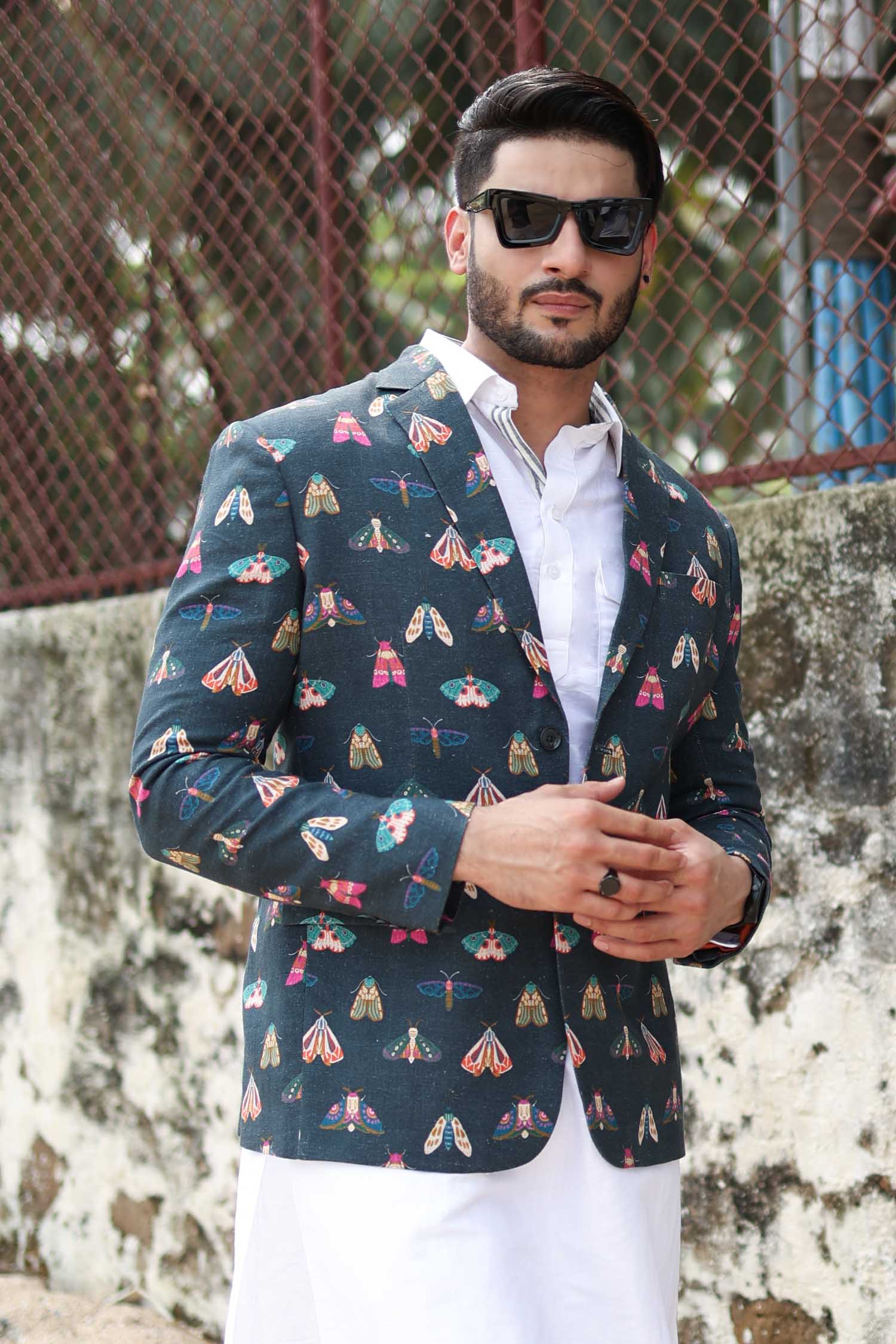 Man wearing a butterfly printed blazer on a wall background a white long shirt, and white trousers. Accessories include sunglasses and a watch.