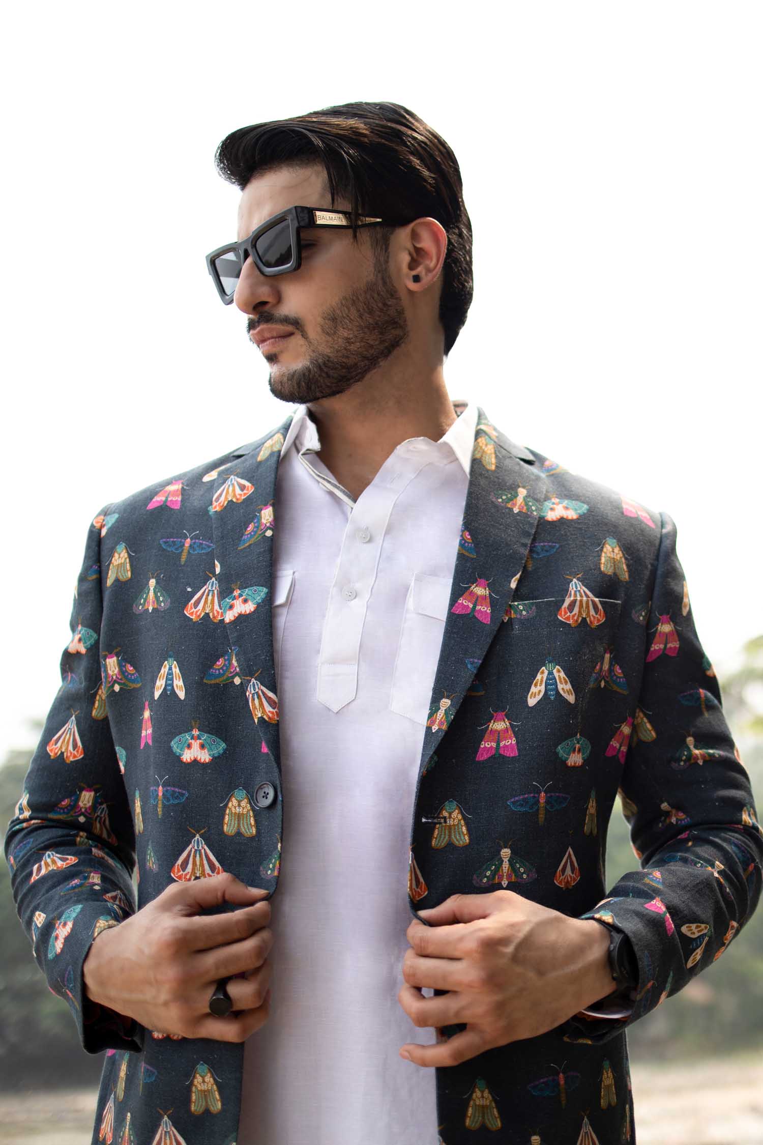 Man wearing a butterfly printed blazer on a wall background a white long shirt, and white trousers. Accessories include sunglasses and a watch.