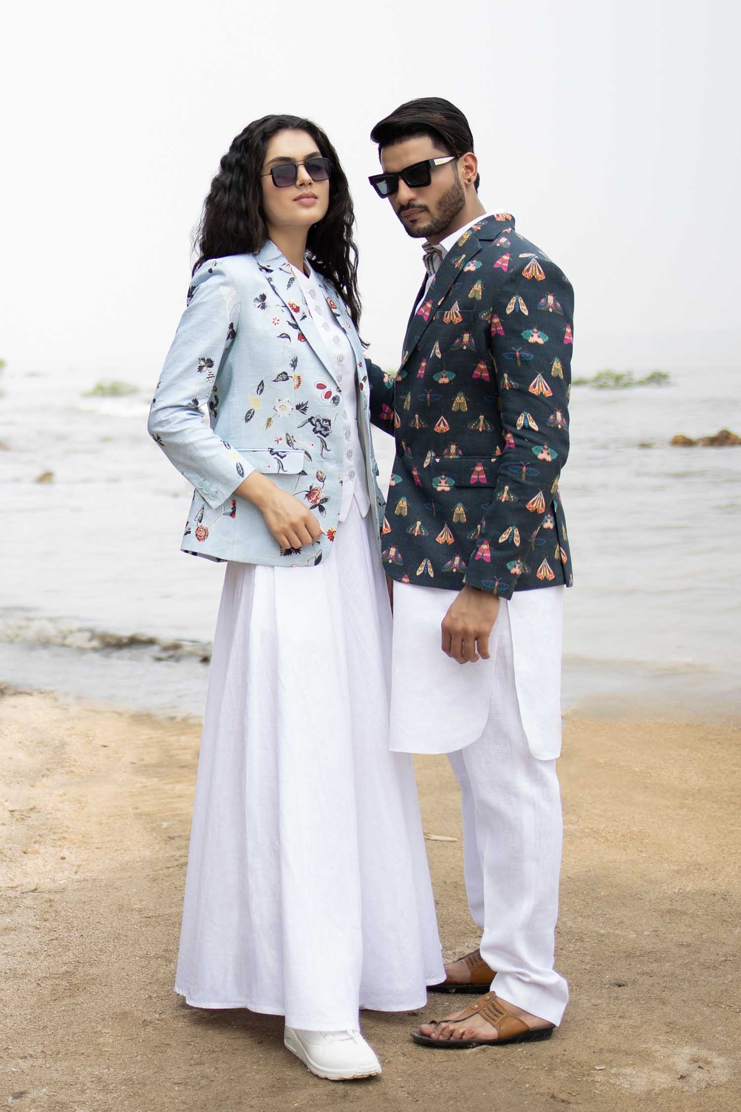 Man wearing a butterfly printed blazer on a wall background a white long shirt, and white trousers. Accessories include sunglasses and a watch.