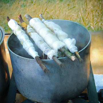 Fabric bundles wrapped tightly around wooden sticks, steaming in a large pot over an open flame