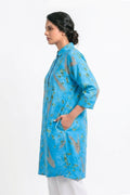 A woman shown in profile wearing a bright blue tunic-style kurta with a subtle gold and green abstract print, paired with white trousers, against a plain white studio background.