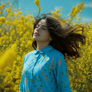 A woman with her hair blowing in the wind stands in a field of bright yellow flowers, wearing a light blue button-up shirt with a subtle floral pattern.