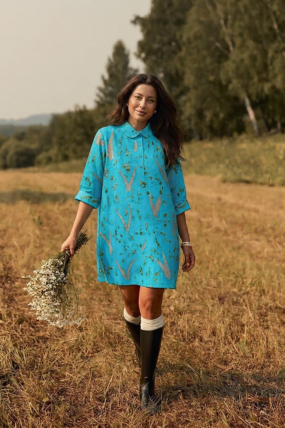 A woman stands in a dry grassy field wearing a turquoise blue patterned dress with three-quarter sleeves and black knee-high boots, holding a small bouquet of white flowers.