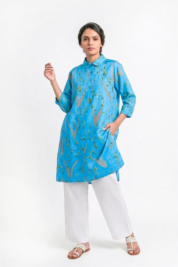 A woman modeling a bright blue kurta with a subtle gold and green floral pattern, paired with white wide-leg trousers and brown sandals, against a plain white studio background.