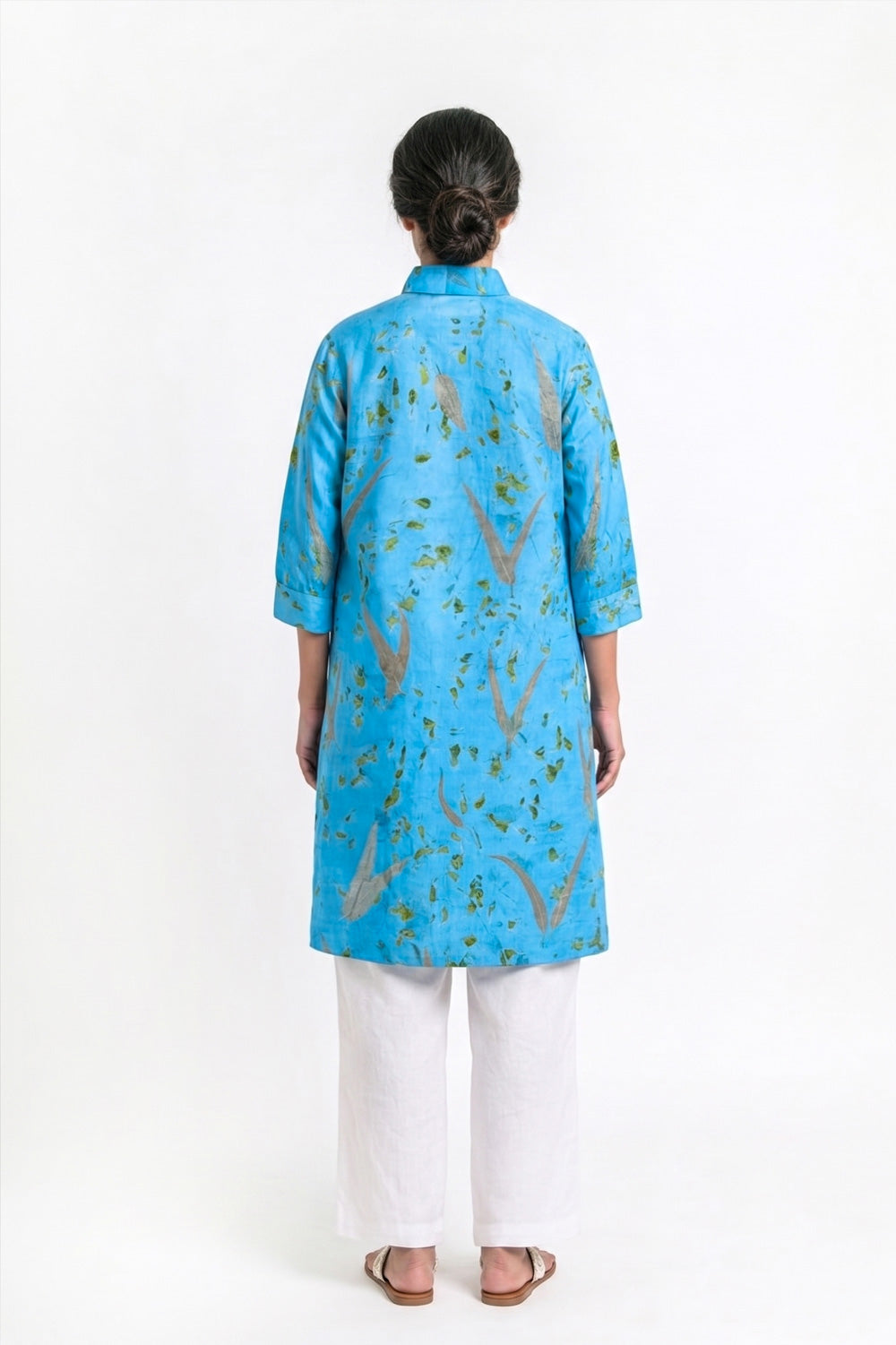 A woman viewed from the back wearing a bright blue tunic with a green and brown abstract pattern, paired with white pants and brown sandals, against a plain white studio background.