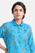 A woman wearing a bright blue short-sleeve button-up shirt with a green and brown abstract leaf-like pattern, photographed against a light gray studio background.