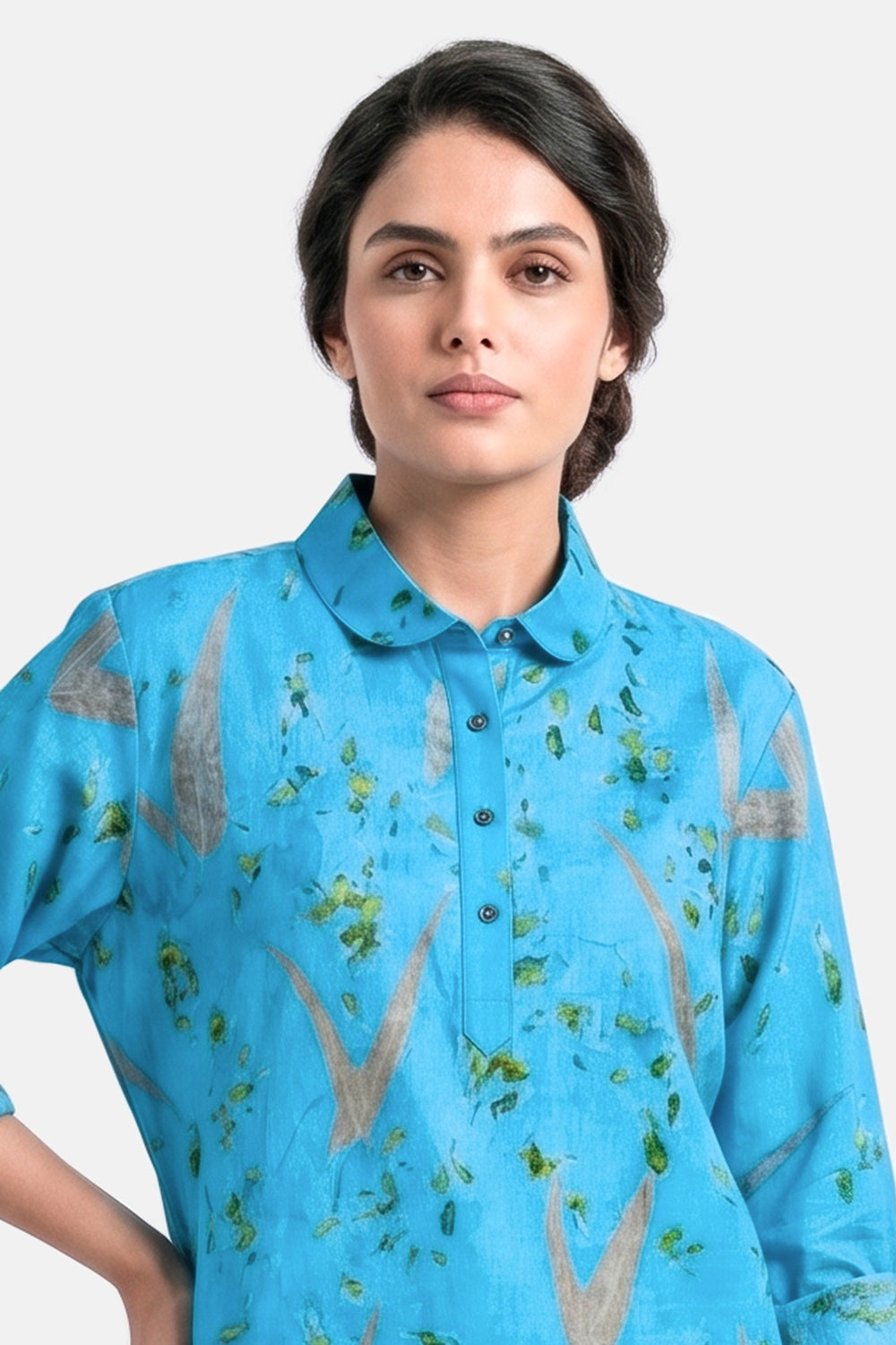 A woman wearing a bright blue short-sleeve button-up shirt with a green and brown abstract leaf-like pattern, photographed against a light gray studio background.