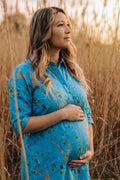 A pregnant woman with blonde-highlighted hair stands in a field of dry golden grass, wearing a bright blue button-up shirt with a green and yellow abstract eco-print pattern.