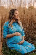 A pregnant woman sits in a field of dry golden reeds, wearing a bright blue button-up shirt dress with a green and beige leaf botanical-print pattern.