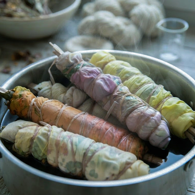 Fabric bundles wrapped around foraged leaves and flowers, tied with string, steaming in a metal pot to transfer natural botanical dye onto the cloth.