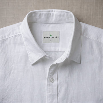 A close-up of a white linen button-down shirt collar with the brand label 'ADAM LEAVES' visible inside the neck, laid flat on a textured beige fabric background.