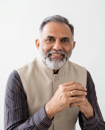 A smiling middle-aged man with gray hair and a beard, wearing a beige mandarin-collar vest over a dark striped button-up shirt, posed against a clean white background.