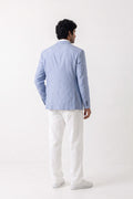 A man shown from the back wearing a light blue linen blazer, white trousers, and white sneakers, standing against a plain white studio background.