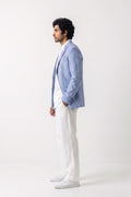 A man in profile wearing a light blue linen blazer over a white shirt and white trousers, standing against a plain white studio background.