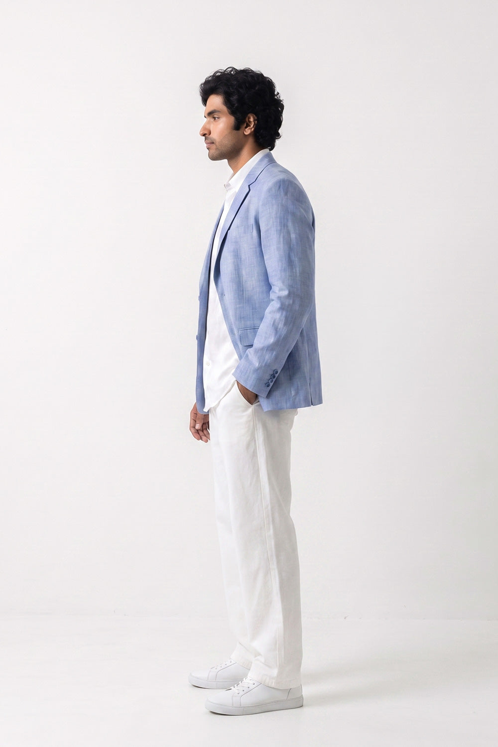 A man in profile wearing a light blue linen blazer over a white shirt and white trousers, standing against a plain white studio background.