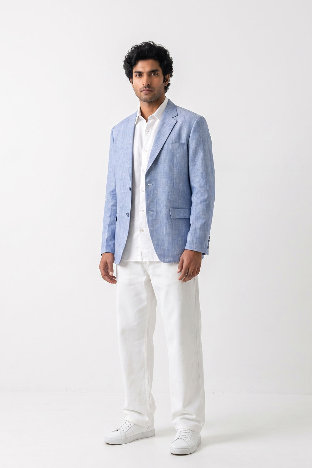 A man modeling a light blue linen-blend blazer over a white shirt and white trousers, standing against a plain white studio background.