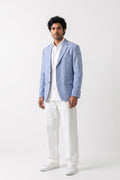 A man modeling a light blue linen-blend blazer over a white shirt and white trousers, standing against a plain white studio background.