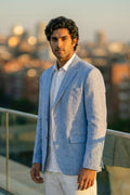 A man wearing a light blue linen blazer over a white shirt and white trousers, standing on a balcony with a blurred city skyline in the background during golden hour.