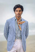 A man wearing a light blue linen blazer, a light blue shirt, a striped orange necktie, and off-white trousers, standing against a soft-focus beach background.