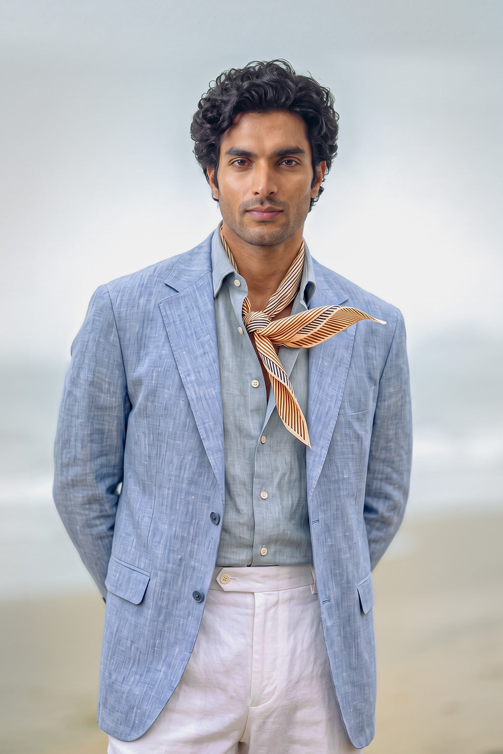 A man wearing a light blue linen blazer, a light blue shirt, a striped orange necktie, and off-white trousers, standing against a soft-focus beach background.