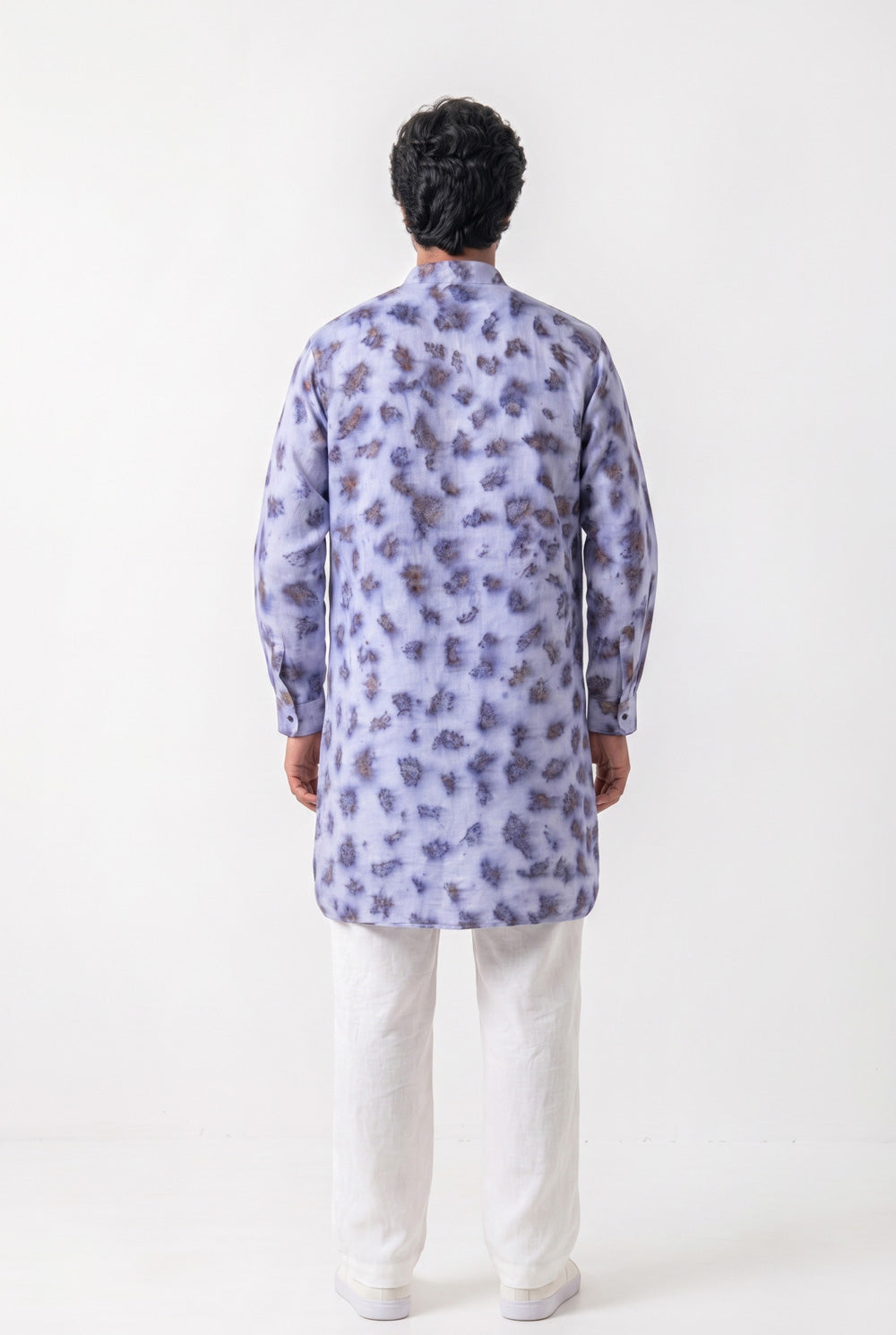 A man viewed from the back wearing a light lavender-blue long kurta with a dark purple tie-dye pattern, paired with white trousers, against a plain white studio background.