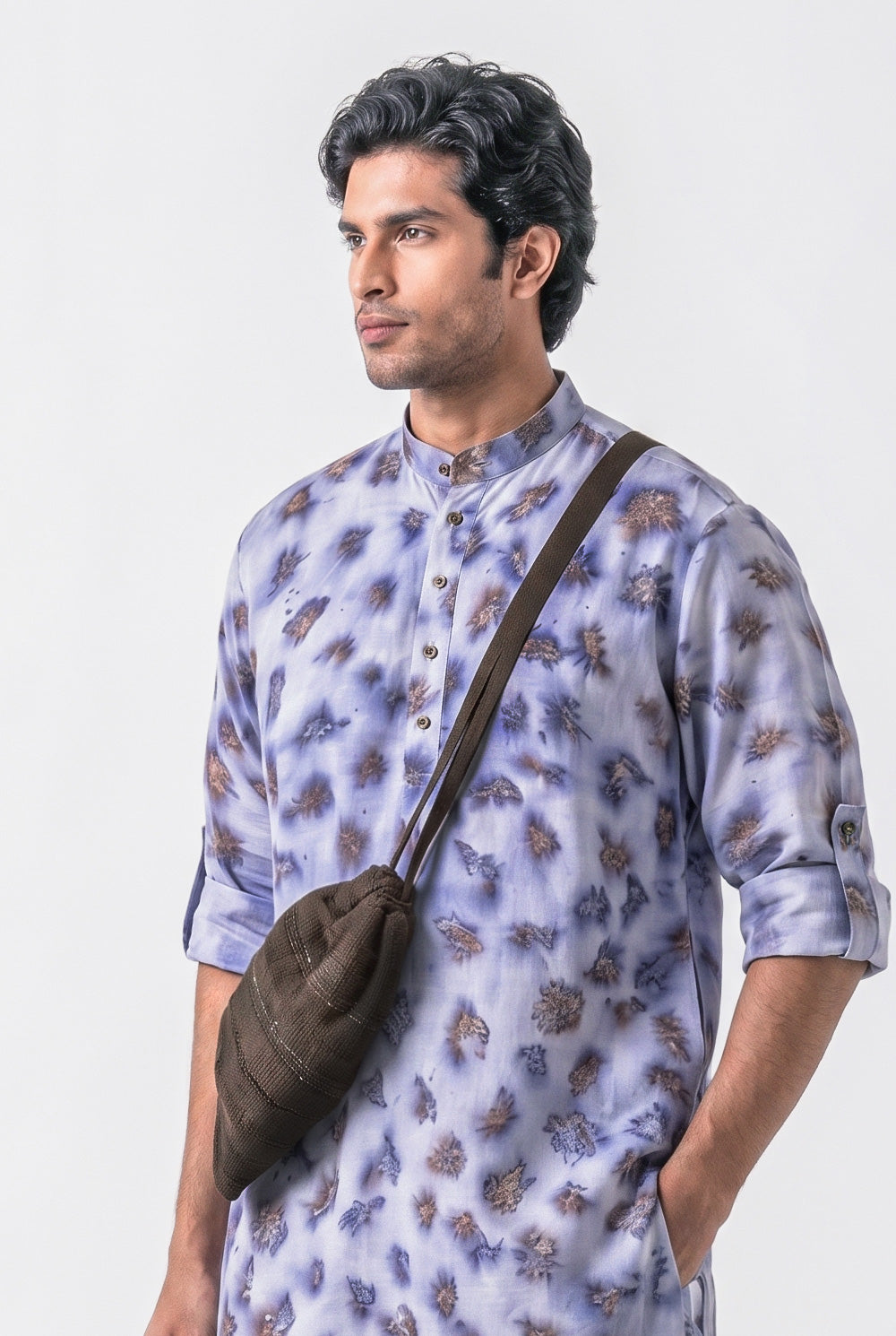 A man wearing a light purple and blue patterned kurta with a brown crossbody bag, photographed against a plain light gray studio background.