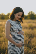 A pregnant woman wearing a sleeveless black and white patterned dress stands in a sunlit field, cradling her baby bump. The background is a soft-focus natural landscape with warm golden light.