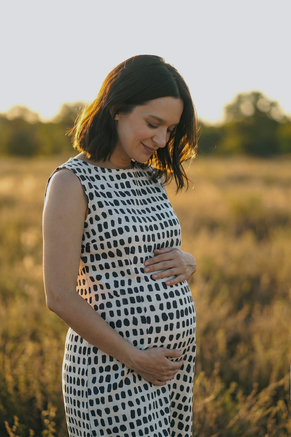 A pregnant woman wearing a sleeveless black and white patterned dress stands in a sunlit field, cradling her baby bump. The background is a soft-focus natural landscape with warm golden light.
