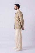 A man shown in profile wearing a beige and white patterned hemp jacket, cream trousers, and white sneakers, standing against a light gray studio background.