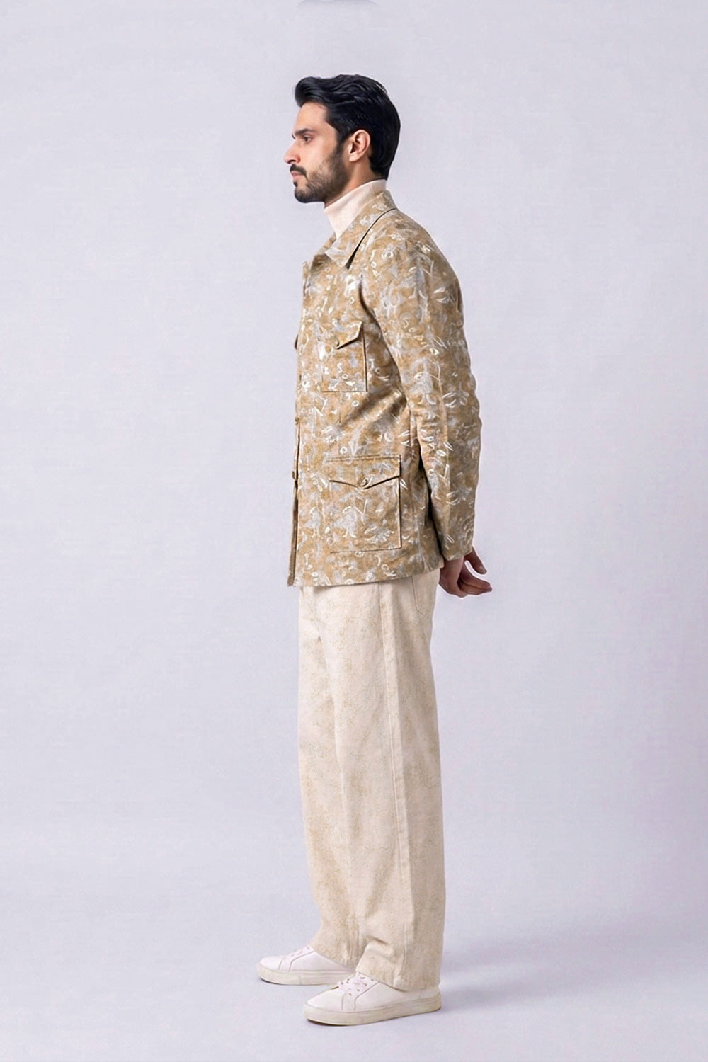 A man shown in profile wearing a beige and white patterned hemp jacket, cream trousers, and white sneakers, standing against a light gray studio background.