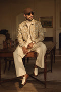 A man wearing a beige and brown patterned hemp jacket, cream turtleneck, cream trousers, brown loafers, and a brown cap sits on a wooden table in a dimly lit room.
