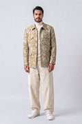 A man shown in front profile wearing a beige and white patterned hemp jacket, cream trousers, and white sneakers, standing against a light gray studio background.
