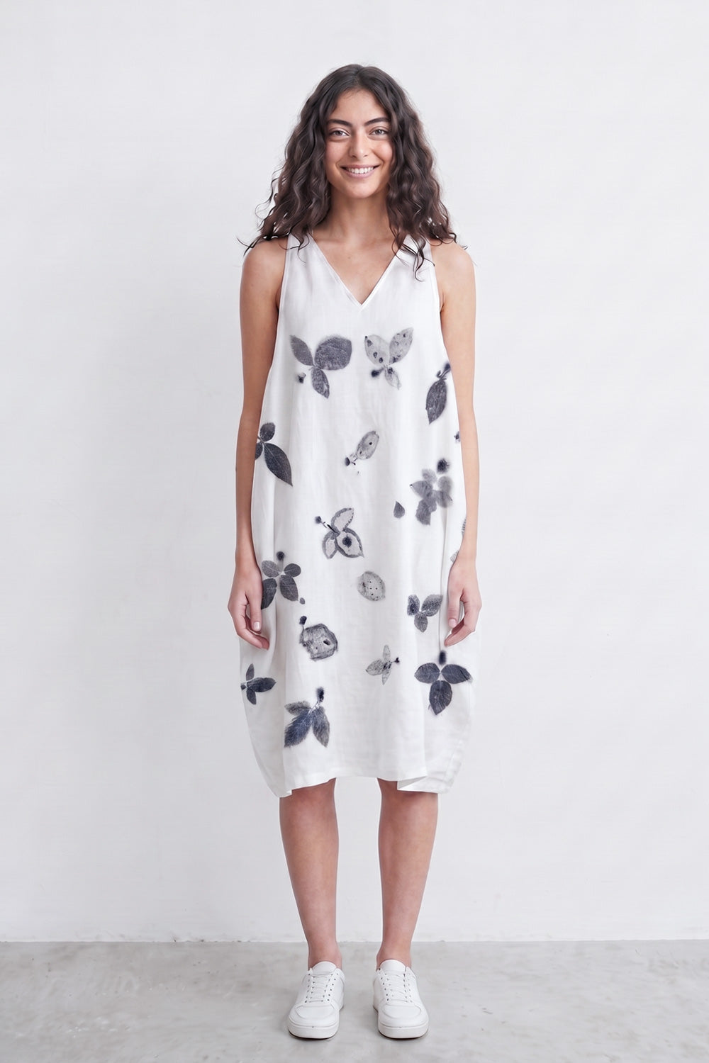 A woman stands against a plain light gray wall wearing a sleeveless white dress with a black and gray botanical print, paired with white sneakers.