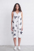 A woman stands against a plain light gray wall wearing a sleeveless white dress with a black and gray botanical print, paired with white sneakers.