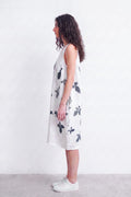 A woman stands in profile wearing a sleeveless white dress with a dark gray botanical leaf print, paired with white sneakers, against a plain white wall.