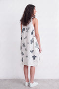 A woman with long curly brown hair is shown from the back wearing a sleeveless white linen dress with a gray botanical and leaf print, paired with white sneakers, against a plain white wall.