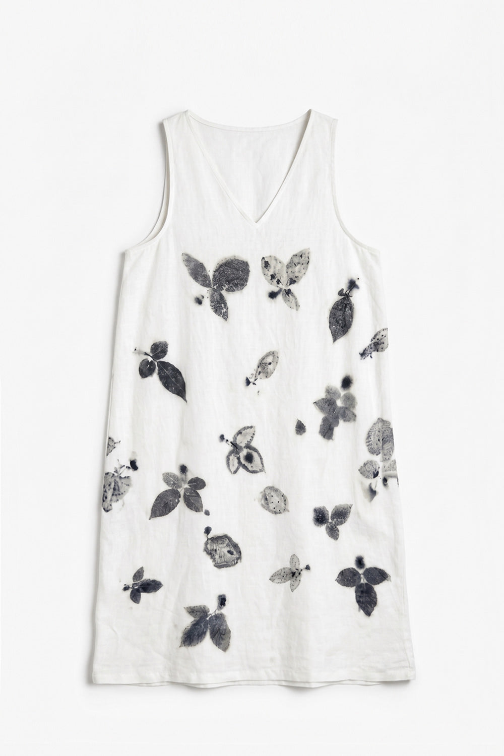 A woman stands against a plain light gray wall wearing a sleeveless white dress with a black and gray botanical print, paired with white sneakers.