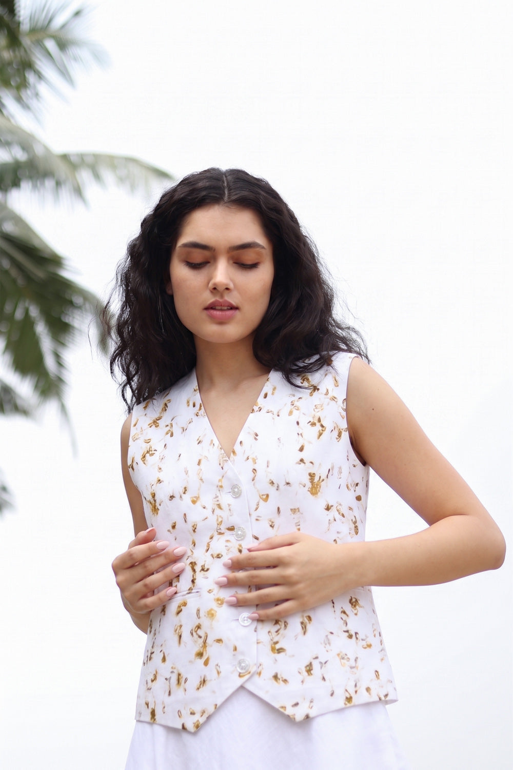 A woman wearing a sleeveless white vest with a gold floral pattern, standing outdoors against a bright white sky with palm leaves in the background.