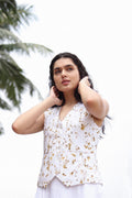 A woman wearing a white sleeveless vest with a gold floral pattern, posing outdoors against a bright white sky with palm leaves in the background.