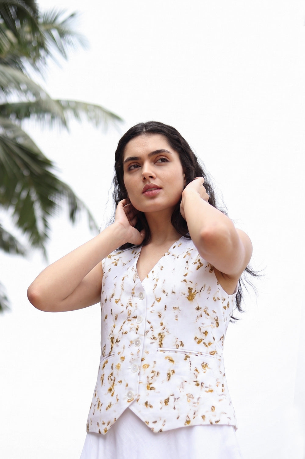 A woman wearing a white sleeveless vest with a gold floral pattern, posing outdoors against a bright white sky with palm leaves in the background.
