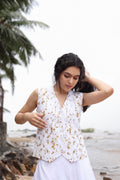 A woman wearing a sleeveless white vest with a gold floral pattern and a white skirt, standing on a beach with palm trees and rocks in the background.
